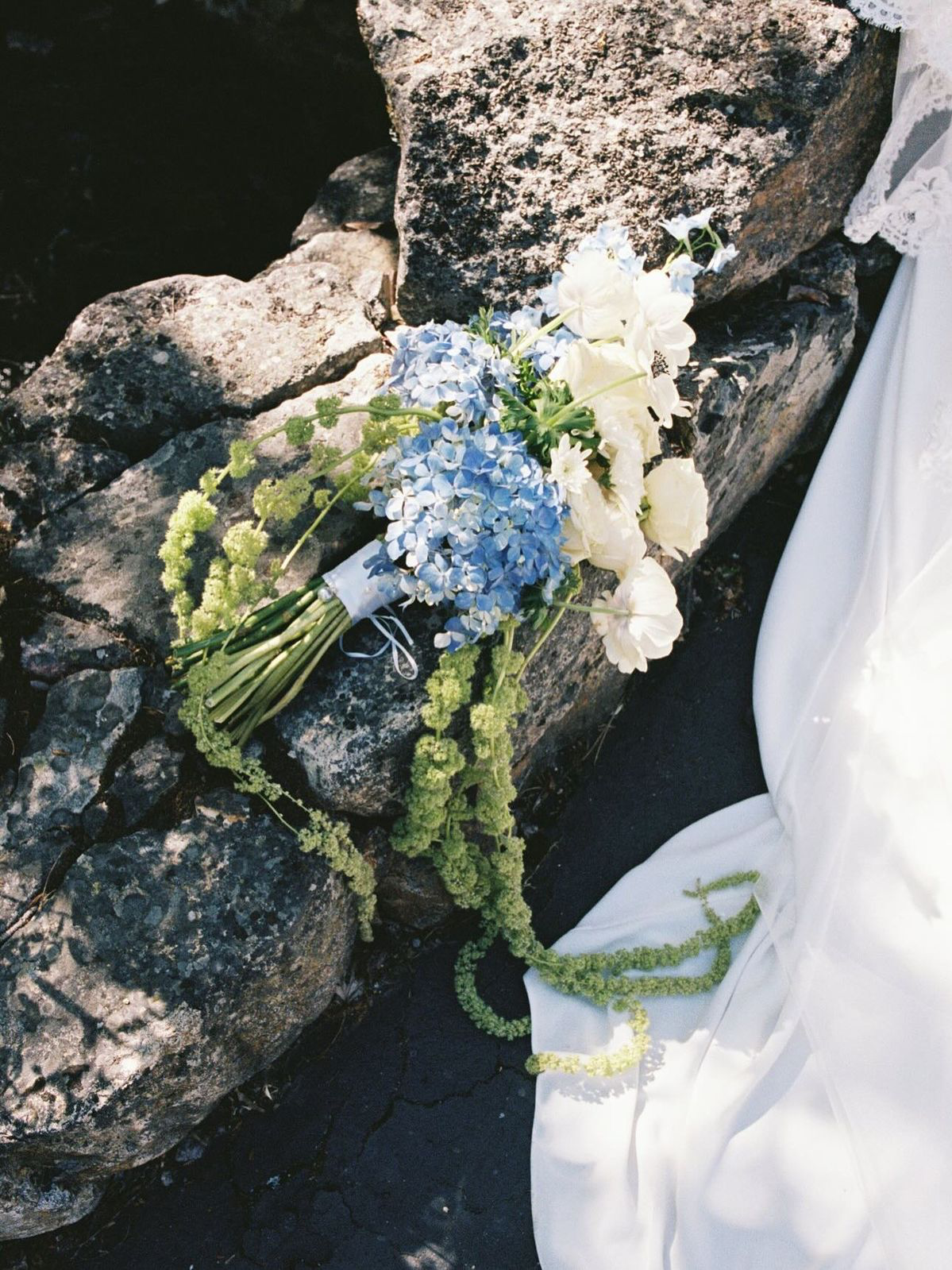 A bridal bouquet of blue hydrangeas and white anemones resting on stone beside a lace wedding dress
